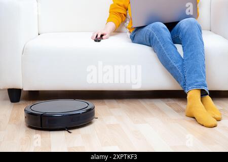 Personne assise sur un canapé blanc avec un ordinateur portable de travail et à distance, tandis qu'un aspirateur robot nettoyage du sol Banque D'Images