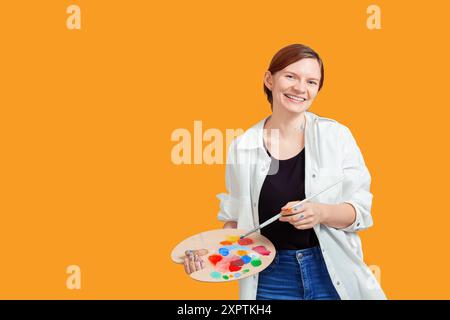 Femme souriante dans une chemise blanche et un Jean tenant une palette de peinture et un pinceau, debout sur un fond orange solide Banque D'Images