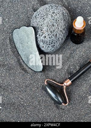 Rouleau de pierre de Gua Sha et de jade avec bouteille d'huile essentielle sur fond de sable noir Banque D'Images