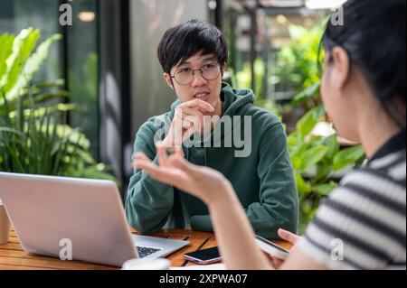 Un jeune homme asiatique dans un sweat à capuche vert parle, discute du travail avec son ami tout en étant assis à une table à l'extérieur. brainstorming, conseil, travail d'équipe Banque D'Images