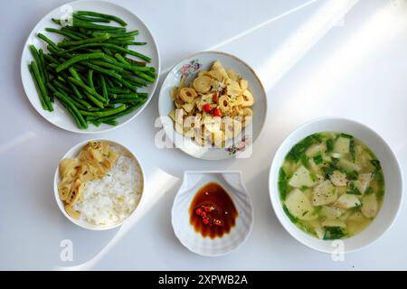 Repas vietnamien avec riz vapeur, tofu et soupe de légumes, haricots verts sautés, pousses de bambou et sauce Chili sur une table blanche, illuminée par na Banque D'Images