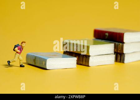 Miniature Tiny People jouet photographie. Un garçon avec un sac à dos d'école debout au-dessus de l'escalier de livre. Isolé sur fond jaune. Photo d'image Banque D'Images