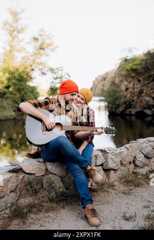 un gars dans un chapeau brillant joue de la guitare avec une fille sur un fond de rochers de granit et un rivière Banque D'Images