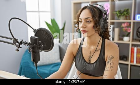 Une jeune femme concentrée avec des écouteurs parle dans un microphone dans un studio de podcast bien éclairé. Banque D'Images