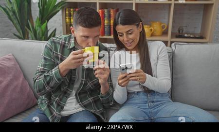 Un couple se détend sur un canapé avec la femme à l'aide d'un smartphone et l'homme sirotant un café dans un salon confortable. Banque D'Images