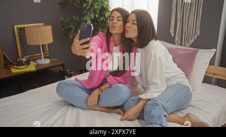 Deux femmes hispaniques s'assoient sur un lit dans une chambre confortable, prenant un selfie avec un smartphone, entouré d'un décor intérieur élégant, y compris un macram Banque D'Images