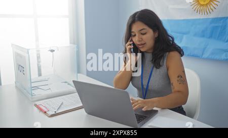 Jeune femme hispanique dans une salle électorale argentine travaillant sur un ordinateur portable à côté d'une urne, parlant au téléphone avec un drapeau argentin derrière Banque D'Images