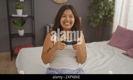 Une jeune femme hispanique heureuse aux cheveux bruns est assise sur un lit dans une chambre confortable à la maison, tenant une manette de jeu et souriant joyeusement. Banque D'Images