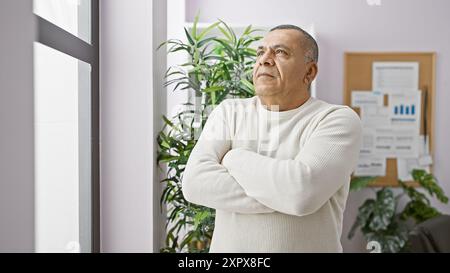 Un homme hispanique contemplatif d'âge moyen se tient debout les bras croisés dans un cadre de bureau moderne. Banque D'Images