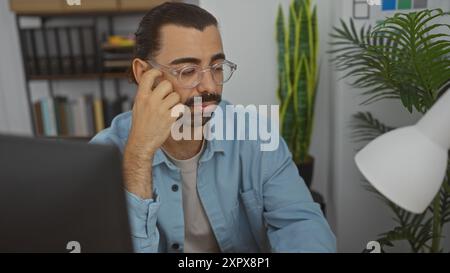 Beau jeune homme hispanique avec une moustache travaillant dans un bureau intérieur, entouré de plantes et d'étagères, portant des lunettes et un shi bleu Banque D'Images
