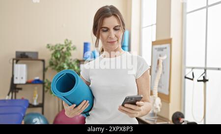 Une femme mature tient un tapis de yoga et un smartphone dans un centre de physiothérapie, représentant le bien-être et la technologie à l'intérieur. Banque D'Images