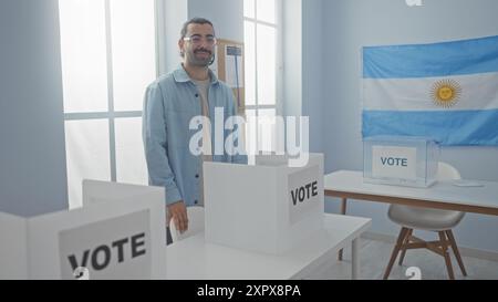 Jeune homme hispanique à l'intérieur d'un collège électoral argentin debout près d'une cabine de vote avec une moustache et portant des lunettes et une chemise en Jean avec le Banque D'Images