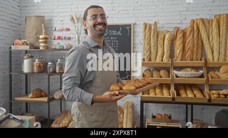 Jeune homme hispanique avec moustache souriant tout en tenant un plateau de croissants dans un intérieur de boulangerie entouré de différents types de pain Banque D'Images