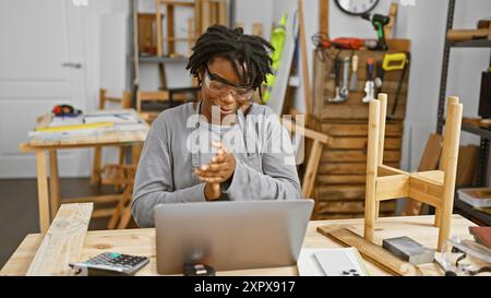 Une jeune femme noire souriante avec des dreadlocks portant des lunettes de sécurité frappe ses mains dans un atelier de menuiserie Banque D'Images