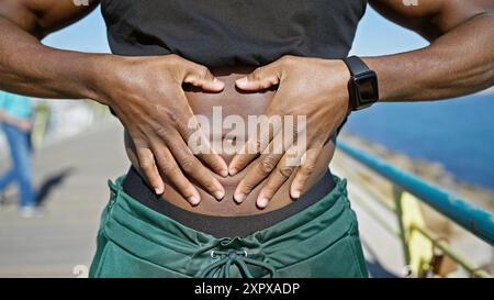 Un homme afro-américain en forme avec une smartwatch repose au bord de la mer, créant une forme de cœur avec ses mains sur son ventre. Banque D'Images