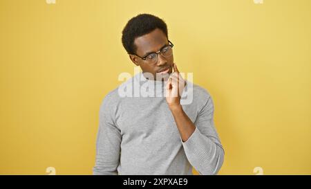 Un jeune homme afro-américain réfléchi portant des lunettes, isolé sur fond jaune, pose pour un portrait. Banque D'Images