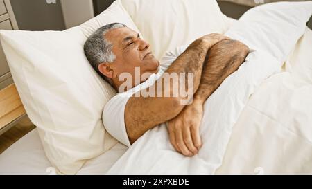 Homme aux cheveux gris dormant paisiblement dans un lit blanc dans un intérieur lumineux de chambre Banque D'Images