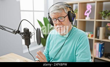 Un homme aux cheveux gris portant des écouteurs parle dans un microphone dans un studio de radio, signalant son professionnalisme et sa concentration. Banque D'Images