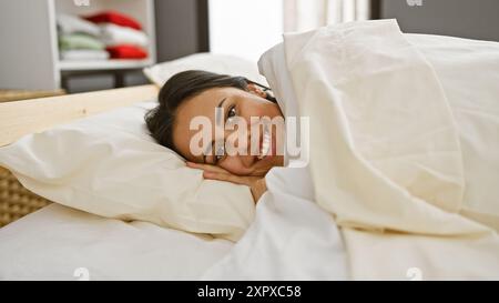 Une jeune femme hispanique souriante se détend confortablement dans une chambre confortable, dégageant un air de chaleur et de tranquillité. Banque D'Images