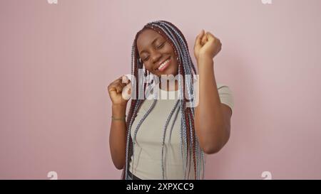 Une joyeuse femme afro-américaine avec des tresses danse sur un fond rose, rayonnant de beauté et de bonheur. Banque D'Images