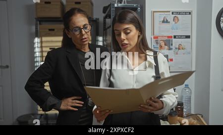 Deux détectives femmes analysent un cas dans le bureau encombré d'un département de police, flanqué d'affiches de personnes disparues. Banque D'Images