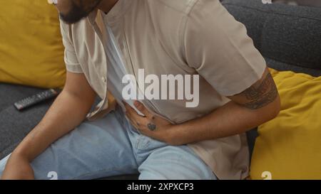 Un homme noir avec un tatouage tient son estomac dans la douleur à la maison, sur un canapé rembourré jaune. Banque D'Images