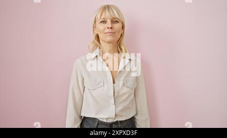 Portrait d'une femme d'âge moyen confiante aux cheveux blonds, habillée de façon décontractée, debout sur un fond rose. Banque D'Images