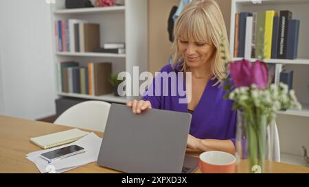 Une femme blonde dans une blouse violette travaillant sur un ordinateur portable à une table en bois dans un bureau à domicile lumineux avec des étagères Banque D'Images