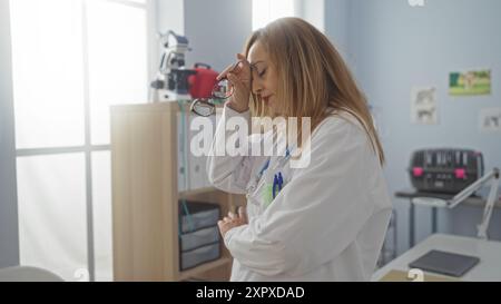 Femme dans une clinique vétérinaire semblant fatiguée tout en tenant ses lunettes, entourée de matériel médical et de paperasse dans une pièce lumineuse et organisée. Banque D'Images