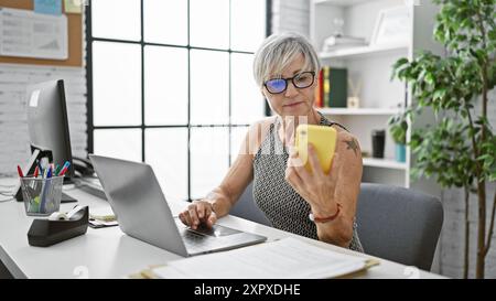 Une femme mature professionnelle utilise un smartphone et un ordinateur portable dans un environnement de bureau, dégageant un multitâche confiant. Banque D'Images