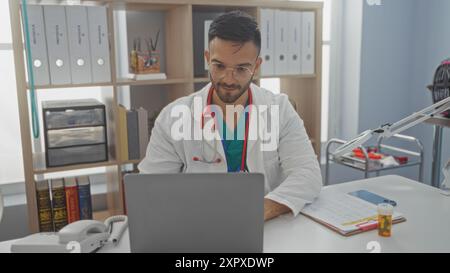 Beau jeune vétérinaire hispanique travaillant sur un ordinateur portable dans un bureau de clinique vétérinaire avec des étagères et du matériel médical en arrière-plan. Banque D'Images