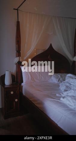 Un lit non fait avec des draps blancs dans une chambre faiblement éclairée avec une tête de lit en bois et une table de chevet avec une lampe. Banque D'Images