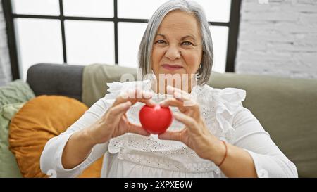 Une femme âgée souriante tient une forme de cœur rouge à la maison, représentant la santé ou l'amour dans un cadre confortable de salon. Banque D'Images