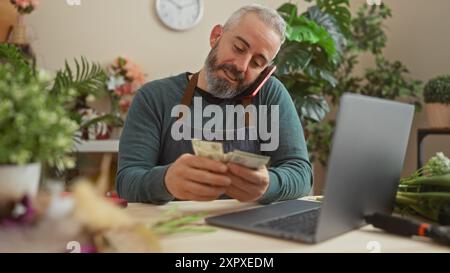 Homme barbu compte de l'argent dans un magasin de fleuriste avec des plantes en arrière-plan, pendant un appel téléphonique en utilisant un ordinateur portable sur le bureau. Banque D'Images