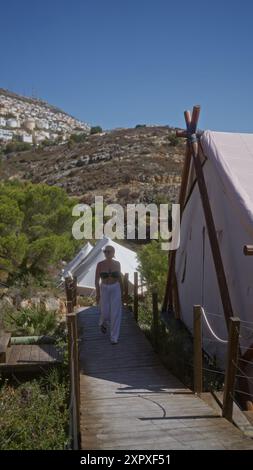 Une jeune femme profitant du plein air se promène le long d’un chemin en bois devant des tentes sur un camping ensoleillé à flanc de colline en afrique. Banque D'Images