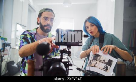 Dans Un studio lumineux, Un jeune homme et une jeune femme caucasienne avec cheveux bleus examinez des séquences vidéo sur Un appareil photo numérique, entouré d'équipements de réalisation de films, incarnant la créativité et la collaboration dans la production cinématographique. Banque D'Images