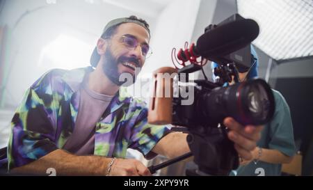 Jeune cinéaste masculin avec barbe et lunettes, souriant tout en utilisant Un appareil photo professionnel dans Un cadre de studio, capturant des séquences pour Un projet de publicité créative. Banque D'Images