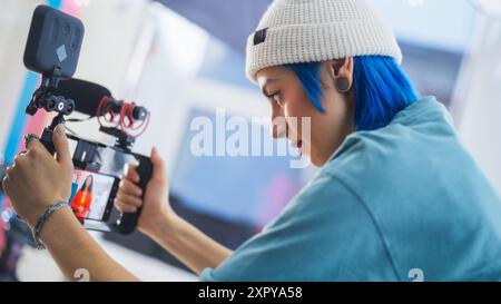 La jeune cinéaste caucasienne focalisée avec cheveux bleus ajuste les paramètres de la caméra sur le plateau de film, capturant un contenu vidéo dynamique et créatif dans Un environnement de studio professionnel. Banque D'Images