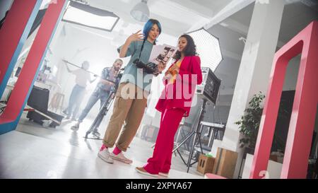 Jeune réalisatrice caucasienne avec Blue Hair discute D'Une scène avec Une actrice noire dans Un costume rose sur Un plateau de film occupé, soulignant le travail d'équipe et la créativité dans Un environnement de production moderne. Banque D'Images