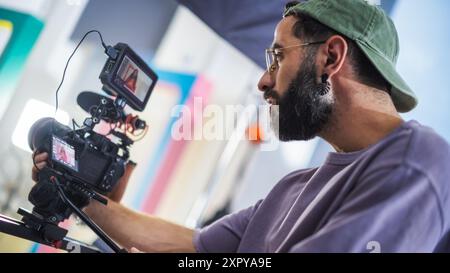 Le directeur de la photographie masculin avec Beard utilise une caméra vidéo professionnelle sur un plateau de tournage, capturant des séquences dynamiques pour Un documentaire ou une émission de télévision, entouré d'un décor coloré. Banque D'Images