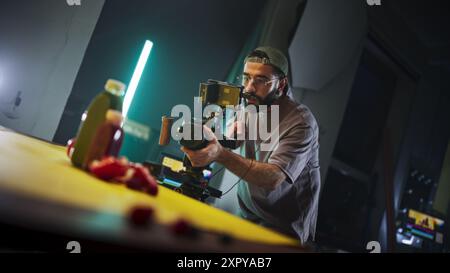 Directeur de la photographie masculin concentré avec barbe, casquette et lunettes, enregistre une scène vibrante sur une caméra professionnelle dans un studio faiblement éclairé, capturant l'essence de la vidéographie moderne et de la narration. Banque D'Images
