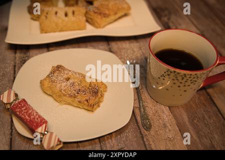 Pâte feuilletée maison remplie de pommes sur une assiette blanche avec une tasse de café, un petit cadeau sur le bord de l'assiette et du sucre en poudre partout Banque D'Images