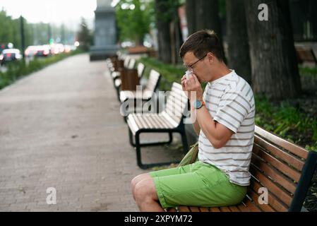 Jeune homme éternuant dans les tissus tout en étant assis sur le banc dans le parc, avec la ligne de bancs et la circulation floue. Les hommes éprouvent une gêne due à l'inflammation et au gonflement du nasopharynx, des allergies saisonnières. Banque D'Images