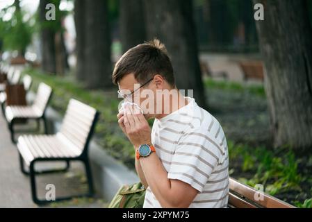 Jeune homme dans des lunettes et une chemise rayée éternuant dans un mouchoir tout en étant assis sur un banc dans un parc. Rhumes, congestion nasale ou allergies saisonnières. Banque D'Images