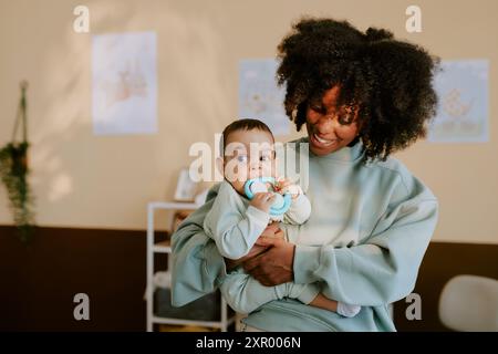 Maman afro-américaine tenant son enfant mâle pendant qu'il mord le hochet bleu Banque D'Images