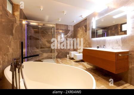 Salle de bains moderne avec murs et sol en marbre. Baignoire au milieu de la pièce. Douche avec parois en verre et lavabo avec un miroir au-dessus. Personne à l'intérieur Banque D'Images