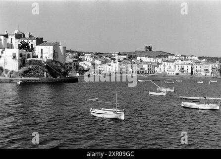 Unbesetzte Boote liegen auf dem Wasser in der Bucht von Cadaqués, im Hintergrund liegen der örtliche Strand und die hellen Häuser des Ortes sowie die ruine des mittelalterlichen Wachturms Torre de sa Riera, auch Torre d'en Reig, Costa Brava 1957. Des bateaux inoccupés se trouvent sur l'eau dans la baie de Cadaqués, avec la plage locale et les maisons de couleur claire de la ville et les ruines de la tour de guet médiévale Torre de sa Riera, également connue sous le nom de Torre d'en Reig, Costa Brava 1957, en arrière-plan. Banque D'Images