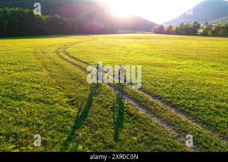 Touristes, couple sur un VTT, cyclistes sur une route de terre à vélo à travers une belle prairie contre le soleil couchant, Dolenjske toplice, Slovénie Banque D'Images