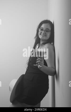 Portrait en noir et blanc d'une femme appuyée contre un mur tenant une tasse de verre avec du vin à l'intérieur. Photo studio. Banque D'Images