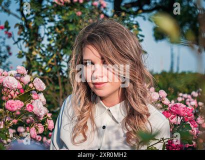 Une jeune femme avec des cheveux coulants est assise au milieu d'un arrangement dense de roses roses en fleurs. Elle regarde directement le spectateur, exsudant un sentiment de joie et t Banque D'Images